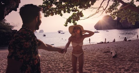 Young travel couple holding hands, woman leading boyfriend. while on Railay Beach at sunset with sun flare. Slow motion, hand held, pov travel concept. Shot on red.