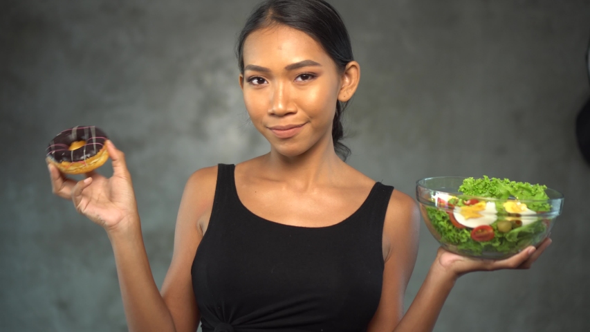 Portrait of beautiful young smiling woman in black t-shirt holding donut and bowl with healthy salad over concrete background. Pretty young woman choosing between healthy and unhealthy food