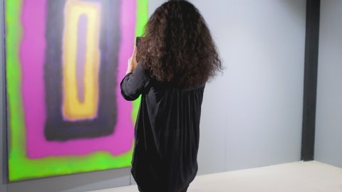 Young woman is photographing abstract artwork in modern gallery. She is using camera of her gadget, focussing on canvas