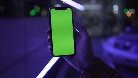 NEW YORK - April 19, 2019: At night light young man hands holding use iPhone with vertical green screen on night city colorful background 