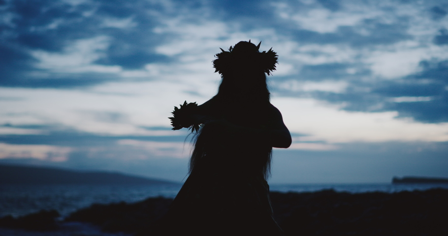 Silhouette of a traditional Hawaiian hula dancer woman dancing on a rugged island landscape at dusk in slow motion with an ocean background