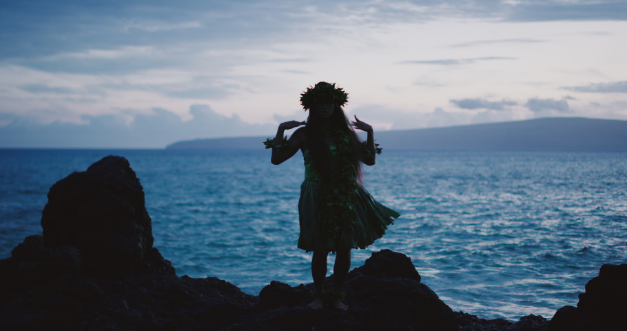 Silhouette of a traditional Hawaiian hula dancer woman dancing on a rugged island landscape at dusk in slow motion with an ocean background