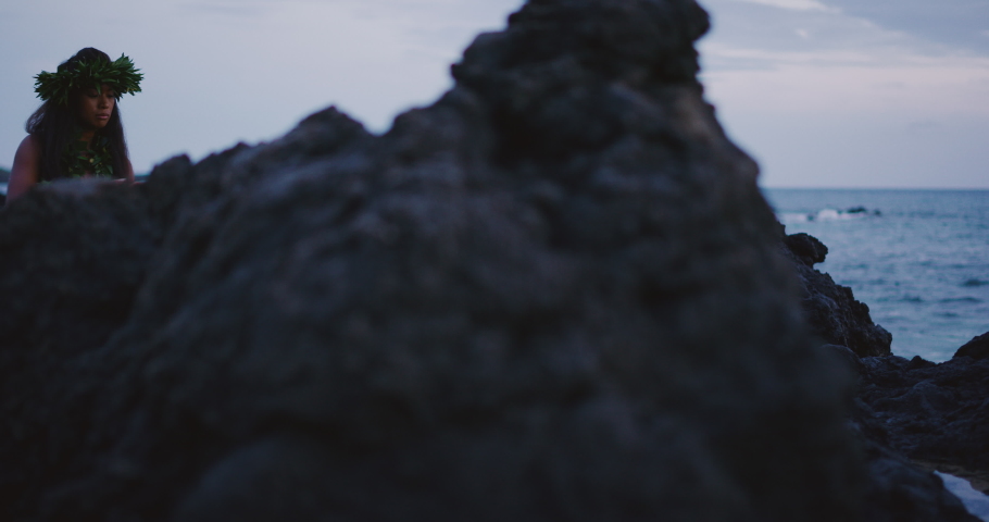 Traditional Hawaiian hula dancing at sunset in slow motion, woman performing Hawaiian hula with haku leis and ti leaf skirt with the ocean in the background