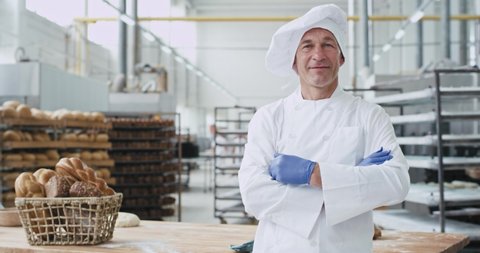 Good looking mature man baker in a big bakery industry smiling large and showing a big like in front of the camera background industrial machine