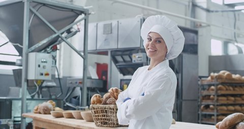 Beautiful and charismatic woman baker in a stylish uniform smiling large in front of the camera in a big bakery industry