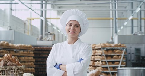In front of the camera charismatic baker woman in a industrial kitchen smiling large and looking stray to the camera background big industrial bakery machine