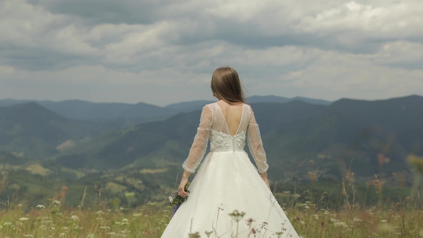Beautiful and lovely bride in wedding dress stay on the mountains with bouquet of flowers. Raises her hands up. Pretty and well-groomed woman. Wedding day. Slow motion