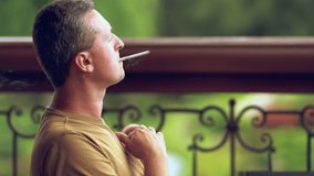 Man enjoying a cigarette outdoors in a park puffing on it before exhaling through his nose with a bannister and greenery behind
