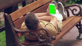 Man relaxing stretched out on a park bench in the sunshine reading on his mobile phone with focus to the blank screen