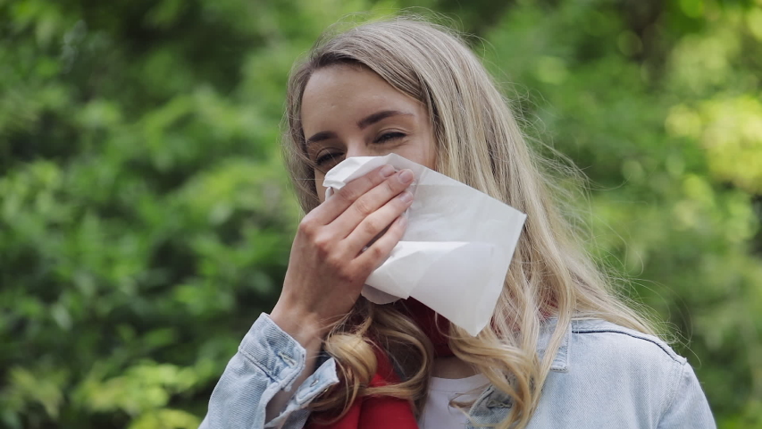 Young woman with with allergy symptom blowing nose standing in the park. Sick girl sneezing and blowing her nose into tissue due to allergy to tree pollen. Royalty-Free Stock Footage #1033097825