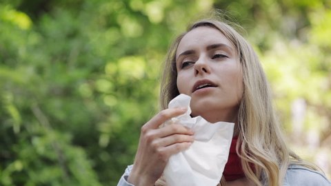 Young woman with with allergy symptom blowing nose standing in the park. Sick girl sneezing and blowing her nose into tissue due to allergy to tree pollen.
