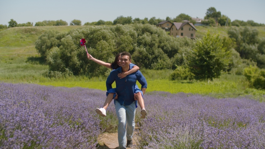 Excited african american man giving piggyback ride to joyful woman with pinwheel and running through lavender bushes. Carefree playful mixed race couple having fun in summer nature in countryside.