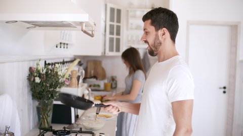 Handheld shot of bearded man in white shirt tossing pancake in pan, then looking at camera with smile while making breakfast in morning. Woman in sleepwear cutting vegetables in background