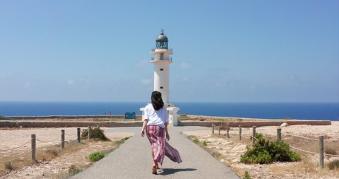 Slow motion of young woman tourist is going towards the Lighthouse near Pilar de la Mola in Formentera Island in Spain during summer holidays time.