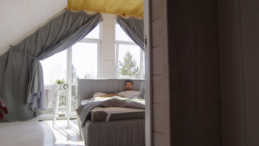 Tracking shot of young woman in pajama running into bedroom and lying down on bed next to her boyfriend with beard. They are laughing and kissing