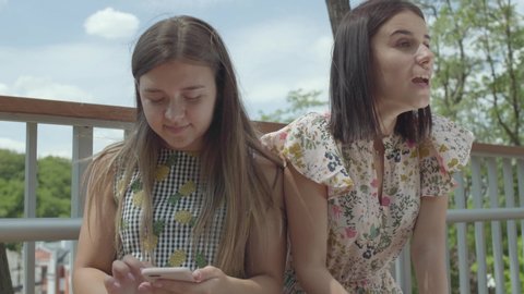 Emotional young woman telling her story to her friend, but she does not listen, texting on the cellphone sitting outdoors. Cute student sharing the problem with her friend, actively gesturing
