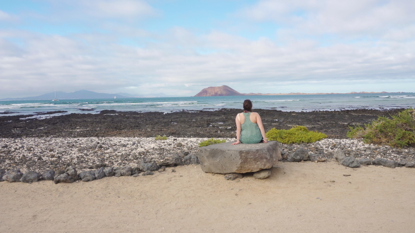 Girl sitting on a rock in Fuerteventura, Spain, contemplating the view of the volcanic islands called Isla de Lobos.