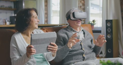 Portrait of elderly couple using tablet computer and virtual reality glasses sitting in living room. Senior and retired people using modern technologies