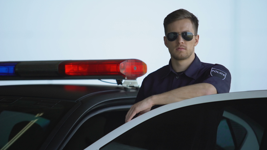 Friendly policeman in sunglasses standing near car and smiling, law and order