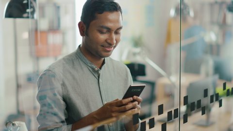Portrait of Handsome Professional Indian Man Uses Mobile Phone, Writes Important Email. Successful Man Using Smartphone Working in Bright Diverse Office