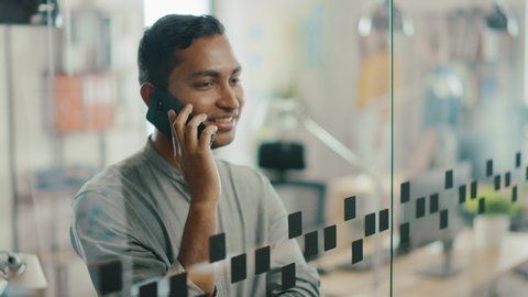 Portrait of Handsome Professional Indian Man Uses Mobile Phone, Talks with Clients and Closes Business Deals. Successful Man Using Smartphone Working in Bright Diverse Office