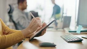 Anonymous Young Woman Sitting at Her Desk She's Drawing, Writing and Using Pen with Digital Tablet Computer. Focus on Hands with Pen. Bright Office where Diverse Team of Young Professionals Work - Powered by Shutterstock - Get 15% off with code: PIKWIZARD15