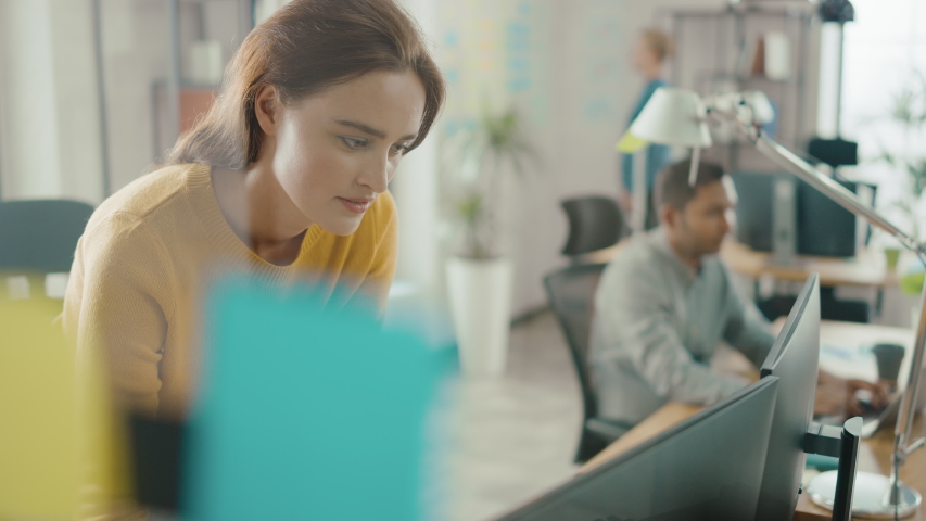 Beautiful and Creative Young Woman Sitting at Her Desk Using Laptop Computer. In the Background Bright Office where Diverse Team of Young Professionals Work Royalty-Free Stock Footage #1033113479