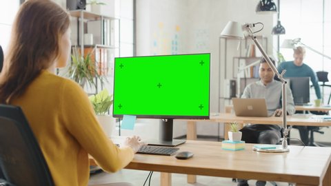 Over the Shoulder: Creative Young Woman Sitting at Her Desk Using Desktop Computer with Mock-up Green Screen. In the Background Bright Office where Diverse Team of Professionals Work on Computers