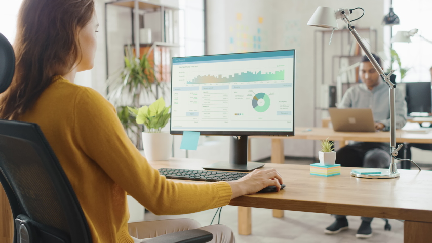 Over the Shoulder: Creative Young Woman Sitting at Her Desk Using Desktop Computer with Screen Showing Project Management, Statistics and Graphs. Office with Diverse Team of Professionals Working - Powered by Shutterstock - Get 15% off with code: PIKWIZARD15