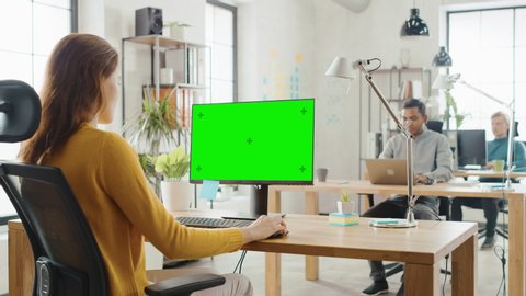 Over the Shoulder:Creative Young Woman Sitting at Her Desk Using Desktop Computer with Mock-up Green Screen. In the Background Bright Office where Diverse Team of Young Professionals Work on Computers
