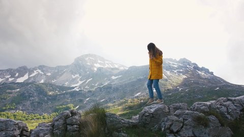 Young cheerful woman in a yellow raincoat walking on rocks with beautiful mountains background, slow motion Video de stock