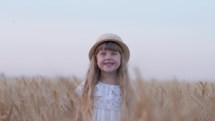 happy childhood, little beautiful fair haired child girl smiles and turns her head standing in golden grain wheat spikes of crop field during weekend outing against sky