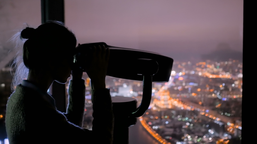 Young woman looking through tourist telescope from skyscraper, exploring night cityscape. Light city street bokeh illumination at evening. Relax, discover and journey concept