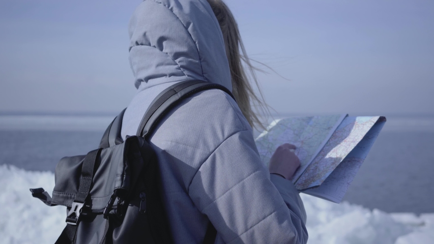 Back view of young blond woman in warm jacket with backpack standing on the glacier checking with the map. Amazing nature of snowy North or South Pole. The tourist in front of the ice blocks