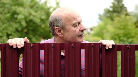 Mature caucasian man carefully watching over the fence. Concept of curious neighbors and private life