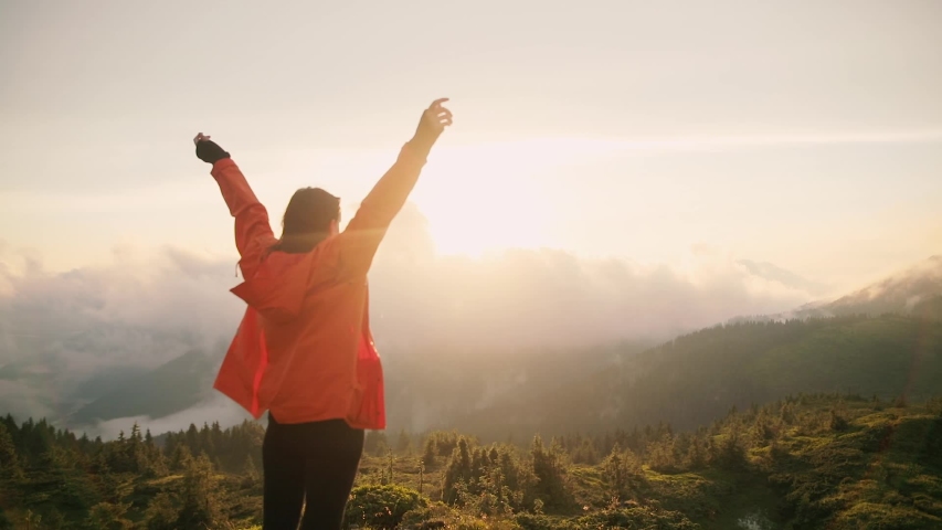 Camera follows hipster millennial young woman in orange jacket running up on top of mountain summit at sunset, jumps on top of rocks, raises arms into air, happy and drunk on life, youth and happiness | Shutterstock HD Video #1033184633