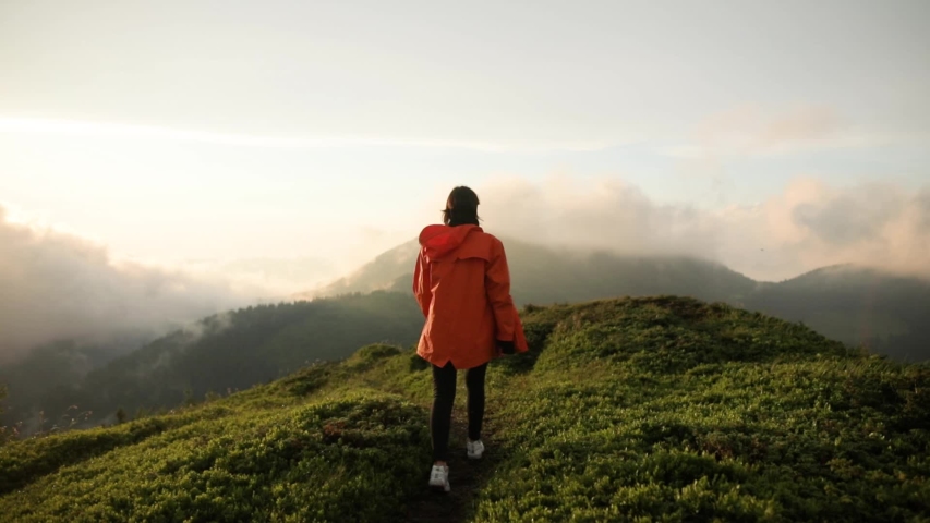 Camera follows hipster millennial young woman in orange jacket running up on top of mountain summit at sunset, jumps on top of rocks, raises arms into air, happy and drunk on life, youth and happiness | Shutterstock HD Video #1033184645