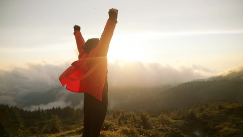 Camera follows hipster millennial young woman in orange jacket running up on top of mountain summit at sunset, jumps on top of rocks, raises arms into air, happy and drunk on life, youth and happiness Stock Video