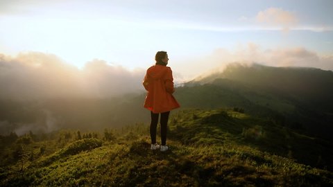 hipster millennial young woman in orange jacke on top of mountain looking at sunset view