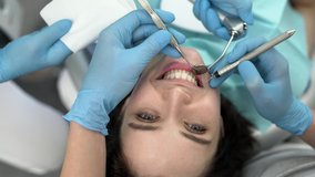 Glad girl in a patient's bib in a dental clinic. Dentist with an assistant are examining her teeth with a help of a dental bur with a mirror and an air polisher. Closeup video recording.
