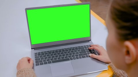 Woman sitting at table and looking at grey laptop computer device with blank green screen in cafe, home or office. Technology, chroma key, template, mockup and entertainment concept