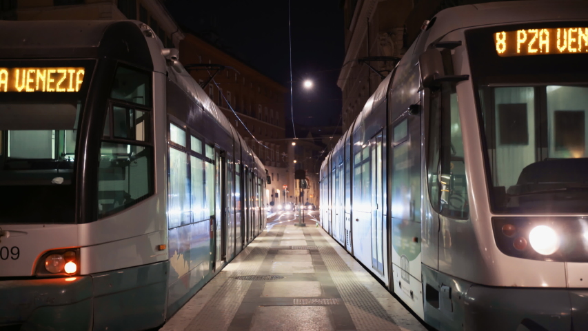 Two traditional italian public trams staying at the railway station on the platforms. One tram having the front lights switched on and the neon inscription above the glass. The doors are opened  