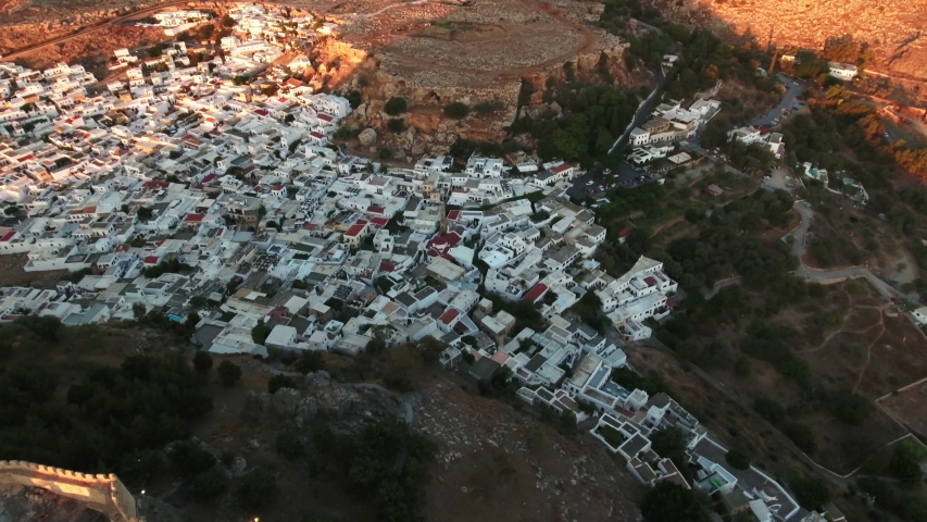 Descending aerial shot of houses with white roofs in Rhodes with trees around the village, and a beautiful landscape, road in the background.