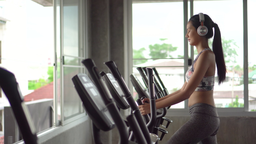 Beautiful young asian woman exercise on elliptical machine and listening music with headphones relaxtion time in fitness gym . sport girl workout , training, healthy lifestyle