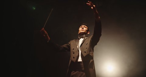 Asian symphony orchestra conductor wearing suit is directing musicians with movement of baton, isolated on black smokey background 4k footage