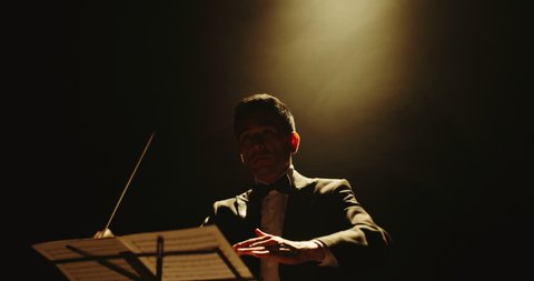 Unrecognizable male symphony orchestra conductor wearing black tux is directing musicians in orchestra pit by moving his hands and baton, studio shot on black background 4k footage