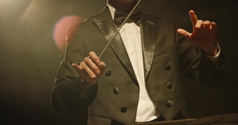 Male orchestra conductor wearing tux standing in front of music stand, controlling musicians by moving his hands and baton. Studio shot on black background 4k footage