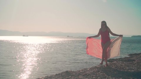 A pregnant girl in a red one-piece swimsuit walks along the seashore in the rays of the setting sun, holding a stole in her hands. Sun overexposure effect.