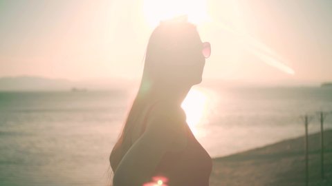 A beautiful young girl in a red one-piece swimsuit and glasses stands on the seashore in the rays of the sunset sun. Sun Overexposure Effect
