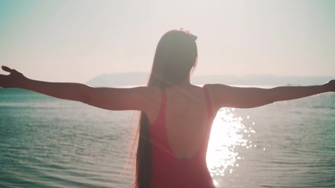 A beautiful young girl in a red one-piece swimsuit stands on the seashore in the sunset sun. A girl stands with her back in the frame, raising her hands up. Sun Overexposure Effect